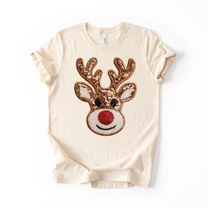 May include: Cream-colored t-shirt featuring a sequined reindeer face. The reindeer has brown antlers, a red nose, and white accents. The design is centered on the front of the short-sleeved shirt, perfect for the holiday season.