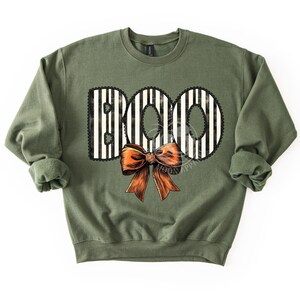 May include: Olive green sweatshirt with the word "BOO" in a black and white striped design, outlined with black lace. An orange bow is centered below the word. The sweatshirt is a casual, long-sleeved garment.