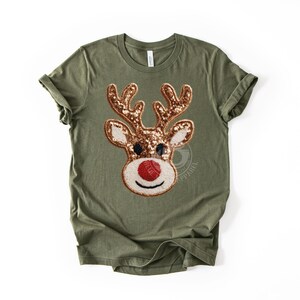 May include: Olive green t-shirt featuring a sequined reindeer face. The reindeer has brown antlers, a red nose, and a white and beige face. The shirt is a crew neck with rolled-up sleeves. The design is centered on the front of the shirt.