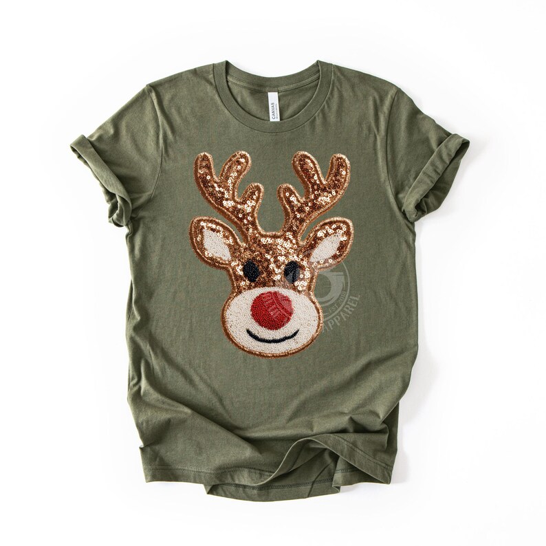 May include: Olive green t-shirt featuring a sequined reindeer face. The reindeer has brown antlers, a red nose, and a white and beige face. The shirt is a crew neck with rolled-up sleeves. The design is centered on the front of the shirt.