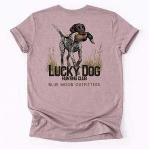 May include: Dusty rose t-shirt featuring a hunting dog carrying a duck in its mouth. The graphic includes the text "LUCKY DOG HUNTING CLUB" and "BLUE MOON OUTFITTERS" in a dark blue color.