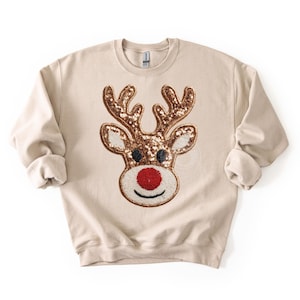 May include: Beige sweatshirt featuring a sequined reindeer face. The reindeer has brown antlers, black eyes, and a red nose. The design is centered on the front of the long-sleeved, crew neck top, perfect for the holiday season.