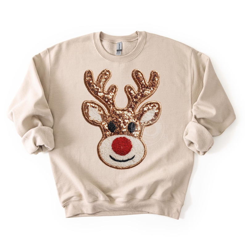 May include: Beige sweatshirt featuring a sequined reindeer face. The reindeer has brown antlers, black eyes, and a red nose. The design is centered on the front of the long-sleeved, crew neck top, perfect for the holiday season.
