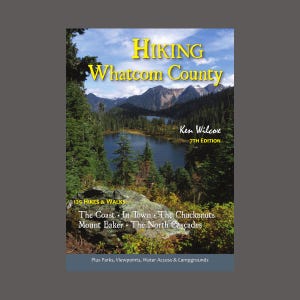 Könnte beinhalten: Buchcover für "Hiking Whatcom County" von Ken Wilcox, 7. Auflage. Das Cover zeigt einen malerischen See, umgeben von Bäumen und Bergen. Das Buch enthält 125 Wanderungen und Spaziergänge in der Gegend, darunter die Küste, in der Stadt, die Chuckanuts, der Mount Baker und die North Cascades. Es enthält auch Informationen über Parks, Aussichtspunkte, Zugang zum Wasser und Campingplätze.