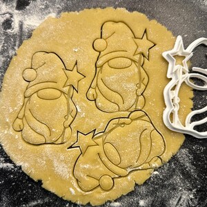 Puede incluir: Primer plano de una masa de galletas redonda y marrón claro con tres formas de gnomos presionadas. Un cortador de galletas de plástico blanco en forma de gnomo está al lado de la masa. Los gnomos tienen varitas de estrella.