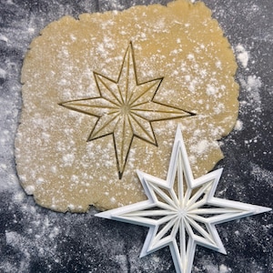 May include: A white star-shaped cookie cutter next to a cut-out star in a piece of dough dusted with flour. The cutter has a detailed, multi-pointed design, perfect for holiday baking.