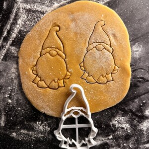 May include: A close-up shot of a cookie dough with two gnome shapes pressed into it. A white plastic gnome-shaped cookie cutter is next to the dough. The dough is dusted with flour, and the background is black.