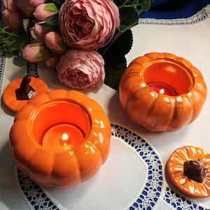 Puede incluir: Dos portavelas de cerámica naranja en forma de calabaza con tapas. Las calabazas tienen una superficie texturizada y están decoradas con tallos marrones. Las tapas tienen un tallo marrón y un pequeño agujero en la parte superior. Las calabazas están iluminadas con velas en su interior.