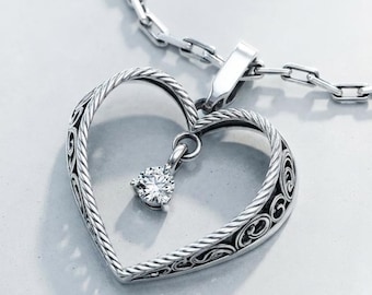 May include: A silver-toned necklace featuring a heart-shaped pendant with a twisted rope-style border and intricate filigree detailing. A single, clear, round-cut gemstone is suspended from the top center of the heart, attached to a rectangular link chain.