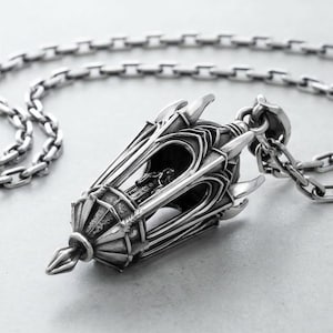 May include: A silver pendant shaped like an ornate lantern with intricate architectural details and pointed accents, suspended from a matching silver link chain. The metal has a polished finish with darkened recessed areas to highlight the design.