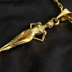 Könnte beinhalten: Eine goldfarbene Halskette mit einem Anhänger in Form einer liegenden Figur. Der Anhänger hängt an einer passenden Goldkette. Die Figur ist detailliert mit Armen, Beinen und Gesichtszügen. Die Kette liegt auf schwarzem Untergrund.
