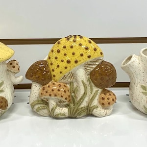 Puede incluir: Un objeto decorativo de cerámica con forma de hongo, con un sombrero amarillo con lunares marrones. El hongo tiene un tallo blanco y varios hongos marrones y beige más pequeños en su base. También hay detalles verdes en forma de hierba.