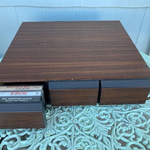 May include: A wooden cassette tape storage unit with three drawers. The drawers are open and contain cassette tapes with titles such as "Beauty and the Beast" and "The Jungle Cats".