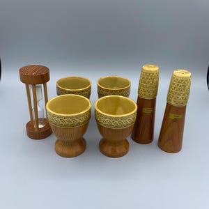 Puede incluir: Colección de utensilios de cocina vintage, que incluye cuatro hueveras, dos saleros y pimenteros y un reloj de arena de madera. Las hueveras tienen un diseño de dos tonos con una base marrón claro y un borde amarillo estampado. Los saleros y pimenteros son marrones con la parte superior amarilla estampada.