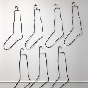 Op de afbeelding: Zeven zwarte metalen sokkenhangers met rood getipte haken zijn tegen een witte muur geplaatst. De hangers hebben de vorm van sokken, met een haak aan de bovenkant om op te hangen. Het ontwerp is minimalistisch en modern.
