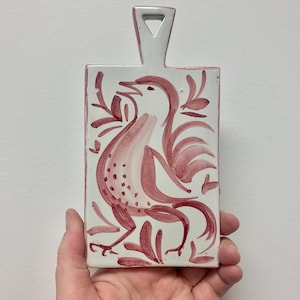 May include: A rectangular, white ceramic chopping board with a handle. The board features a hand-painted design of a bird in shades of red. The bird is surrounded by red floral accents. The handle has a triangular opening.