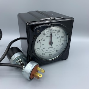 Op de afbeelding: Een zwarte elektrische timer met een wit gezicht en een zwart snoer. De timer heeft een grote wijzerplaat met nummers van 0 tot 90 en een kleinere wijzerplaat met nummers van 0 tot 60. De timer is gemarkeerd met "STANDARD".