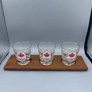 May include: Three clear shot glasses with gold rims, each featuring a red maple leaf and Olympic rings design. The glasses sit on a wooden tray. The text "CANADA 88" is visible on the glasses.