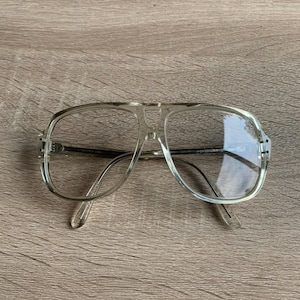 May include: Clear aviator-style spectacles with a light-coloured frame. The spectacles have a double bridge and clear lenses. The temples are also clear. The spectacles are resting on a wooden surface.