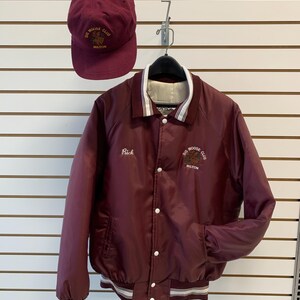 Peut inclure: Une veste et une casquette de baseball bordeaux du Big Moose Club. La veste a des bordures blanches sur le col, les poignets et la taille, avec le nom "Rick" brodé sur la poitrine. La casquette et la veste présentent le logo du club et la ville de Milton.
