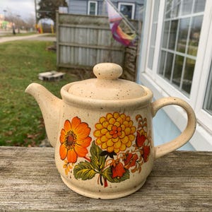 Può includere: Una teiera in ceramica beige con una glassa macchiettata e un design floreale con fiori arancioni, gialli e verdi. La teiera ha un coperchio e un manico.