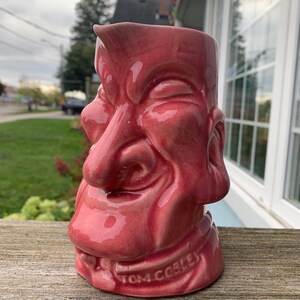 Può includere: Un'anfora in ceramica rosa con un viso scolpito sul davanti. Il viso ha un naso grande e un ampio sorriso. L'anfora è firmata "Tom Copley" sul fondo.