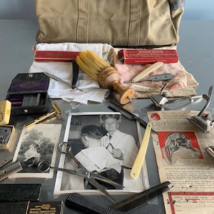 Op de afbeelding: Een verzameling vintage kappersgereedschap en accessoires, waaronder scheermessen, scharen, kammen en een scheerkwast, gerangschikt op een reflecterend oppervlak. Een canvas tas en vintage foto's suggereren een kapperszaak thema.