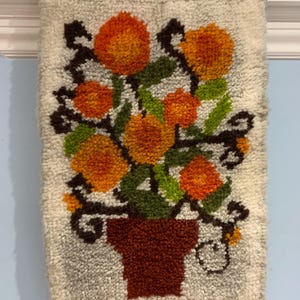 May include: A white wall hanging with a brown pot and orange flowers. The flowers are in a stylized design with green leaves and brown stems.