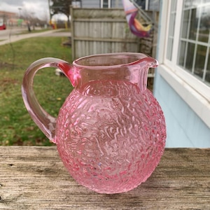 May include: A pink glass pitcher with a textured surface. The pitcher has a round body and a handle.