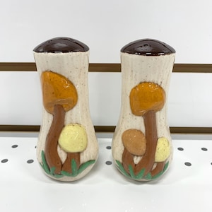 Puede incluir: Un par de saleros y pimenteros de cerámica. Los saleros son de color blanquecino con tapas marrones y presentan diseños de setas en tonos naranja, amarillo y marrón. Las setas tienen hierba verde en la base.