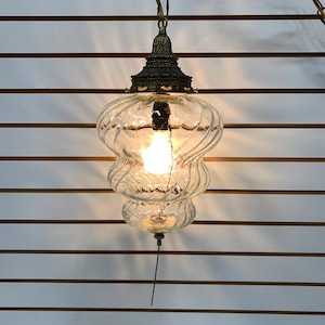 Op de afbeelding: Vintage glazen hanglamp met een wervelend ontwerp. De lamp heeft een messingkleurige bovenkant en ketting, en een trekketting. De gloeilamp brandt en geeft een warme gloed. Een decoratieve verlichtingsarmatuur.