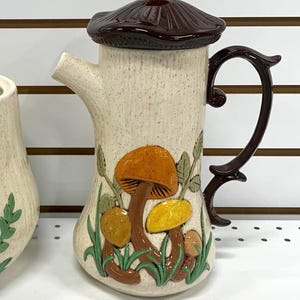 May include: A ceramic teapot with a brown mushroom-shaped lid and handle. The beige body is decorated with colourful mushrooms and green foliage. The spout is on the left side.