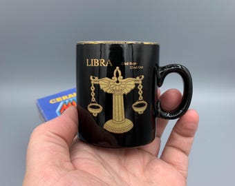 Pode incluir: Caneca de cerâmica preta com borda dourada e um símbolo do zodíaco de Libra em ouro com as datas "23 de setembro - 22 de outubro".