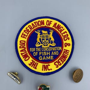 Peut inclure: Ensemble de badges et d'épingles vintage sur le thème de la pêche et de la chasse. Le patch principal est rond, avec le texte "ONTARIO FEDERATION OF ANGLERS & HUNTERS" et un blason. D'autres épingles incluent un canard et une épingle avec le texte "2000".