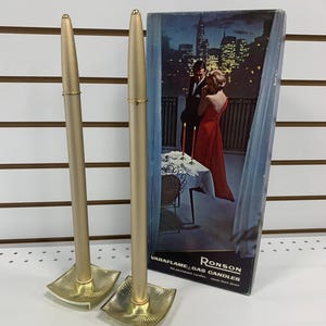May include: Two gold-coloured Ronson Varaflame gas candles with square bases. The candles are tall and slender, with a matching box featuring a couple on a balcony overlooking a city at night. The box reads "Varaflame Gas Candles".