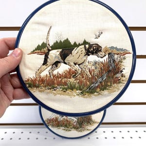 Può includere: Arte murale rotonda raffigurante un cane da caccia in una scena di campo. Il cane è marrone e bianco, con una cornice blu. L'opera d'arte raffigura un cane che corre attraverso l'erba alta, con alberi e uccelli sullo sfondo.