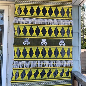 Peut inclure: Une tenture murale tissée avec un motif géométrique. La pièce présente un motif de losanges jaunes et noirs, des franges blanches et des figures blanches sur un fond vert. Les bords supérieur et inférieur ont des bandes décoratives jaunes et noires.