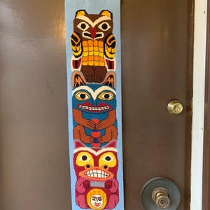 Può includere: Arazzo tessuto con un design a totem colorato. Il totem, su sfondo azzurro, presenta figure di animali nei toni del marrone, giallo, rosso e blu. Appeso a una porta marrone, è un oggetto decorativo.