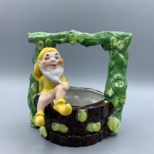 Op de afbeelding: Een keramische plantenbak in de vorm van een put met een kleine kabouter die op de rand zit. De kabouter draagt een geel shirt en bruine laarzen. De put is versierd met groene wijnstokken en bruine stenen.