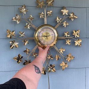 May include: A vintage gold-toned wall clock with a circular face and Roman numerals. The clock is surrounded by a decorative arrangement of stylized leaves and branches. The clock's face is approximately 6 inches in diameter.