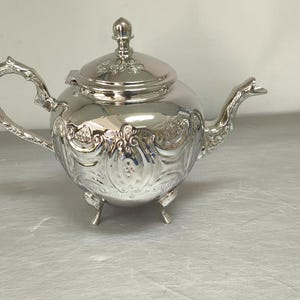 May include: A silver-colored teapot with ornate floral and scrollwork detailing. The teapot has a curved handle, a spout, and a lid with a decorative finial. It sits on three small feet, and reflects the surrounding light.