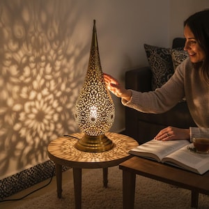 Op de afbeelding: Een druppelvormige tafellamp van antiek messing met ingewikkelde, lasergesneden bloemmotieven. De lamp verspreidt een warme gloed, werpt gedetailleerde, symmetrische schaduwen op de muur en staat op een ronde voet.