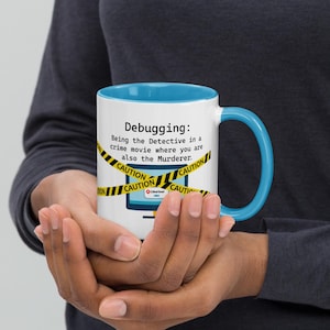 May include: A white coffee mug with a blue rim and handle. The mug features a graphic of a computer screen with caution tape around it. The text on the mug reads "Debugging: Being the Detective in a crime movie where you are also the Murderer."