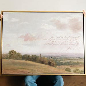 Puede incluir: Una pintura de paisaje enmarcada con un marco dorado. La obra de arte representa una escena pastoral con colinas, árboles y un cielo suave y nublado. El texto "For God so loved the world..." está escrito en el lienzo. La paleta de colores es apagada con tonos terrosos.