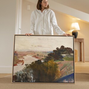 May include: A framed landscape painting depicting a river flowing through a valley. The artwork uses a muted palette of greens, browns, and blues, with a pale sky. The painting is held by a person wearing a white shirt.