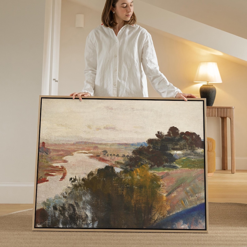 May include: A framed landscape painting depicting a river flowing through a valley. The artwork uses a muted palette of greens, browns, and blues, with a pale sky. The painting is held by a person wearing a white shirt.