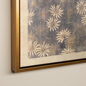 May include: A framed canvas art piece featuring a repeating floral pattern. The flowers are white with tan centers, set against a blue background. The artwork is framed in a gold-colored wooden frame, adding a touch of elegance to the piece.