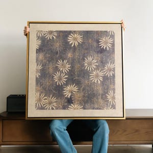 May include: A framed artwork with a repeating pattern of cream-coloured floral shapes on a dark blue background. The artwork is encased in a gold-coloured frame.