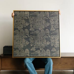 May include: A framed square artwork featuring a dark blue fabric with a repeating pattern of birds and floral elements in a light beige color. The frame is a thin, gold color. The artwork is held up by a person.