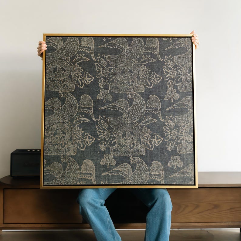 May include: A framed square artwork featuring a dark blue fabric with a repeating pattern of birds and floral elements in a light beige color. The frame is a thin, gold color. The artwork is held up by a person.